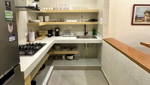 A compact kitchen with a refrigerator, stove, and sink. There are shelves above the counter holding dishes and glasses. - Casa Stephanie- Prívate Pool (Cartagena de Indias)