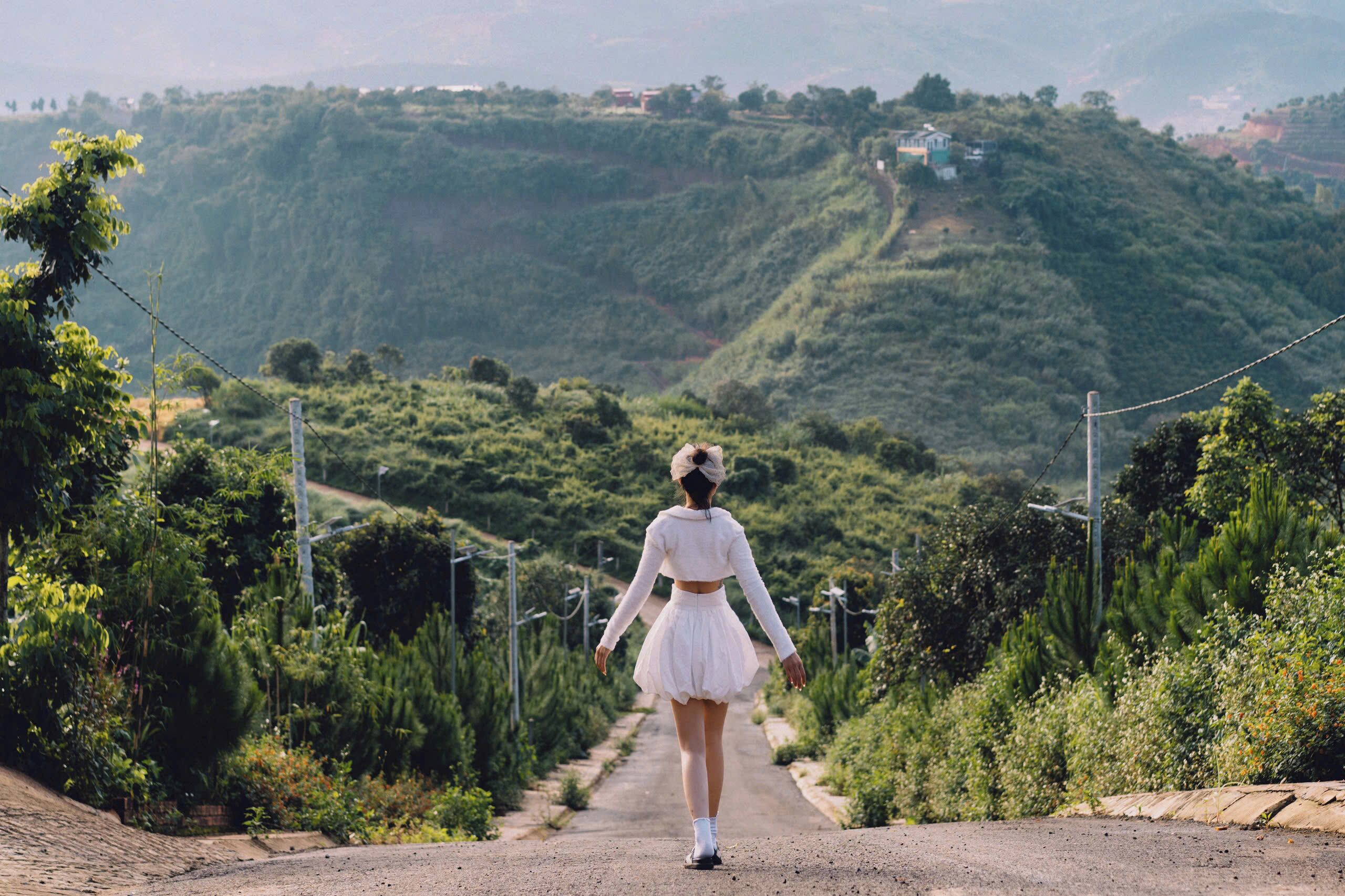 Una mujer con un vestido blanco camina por un camino rural rodeado de vegetación y colinas.