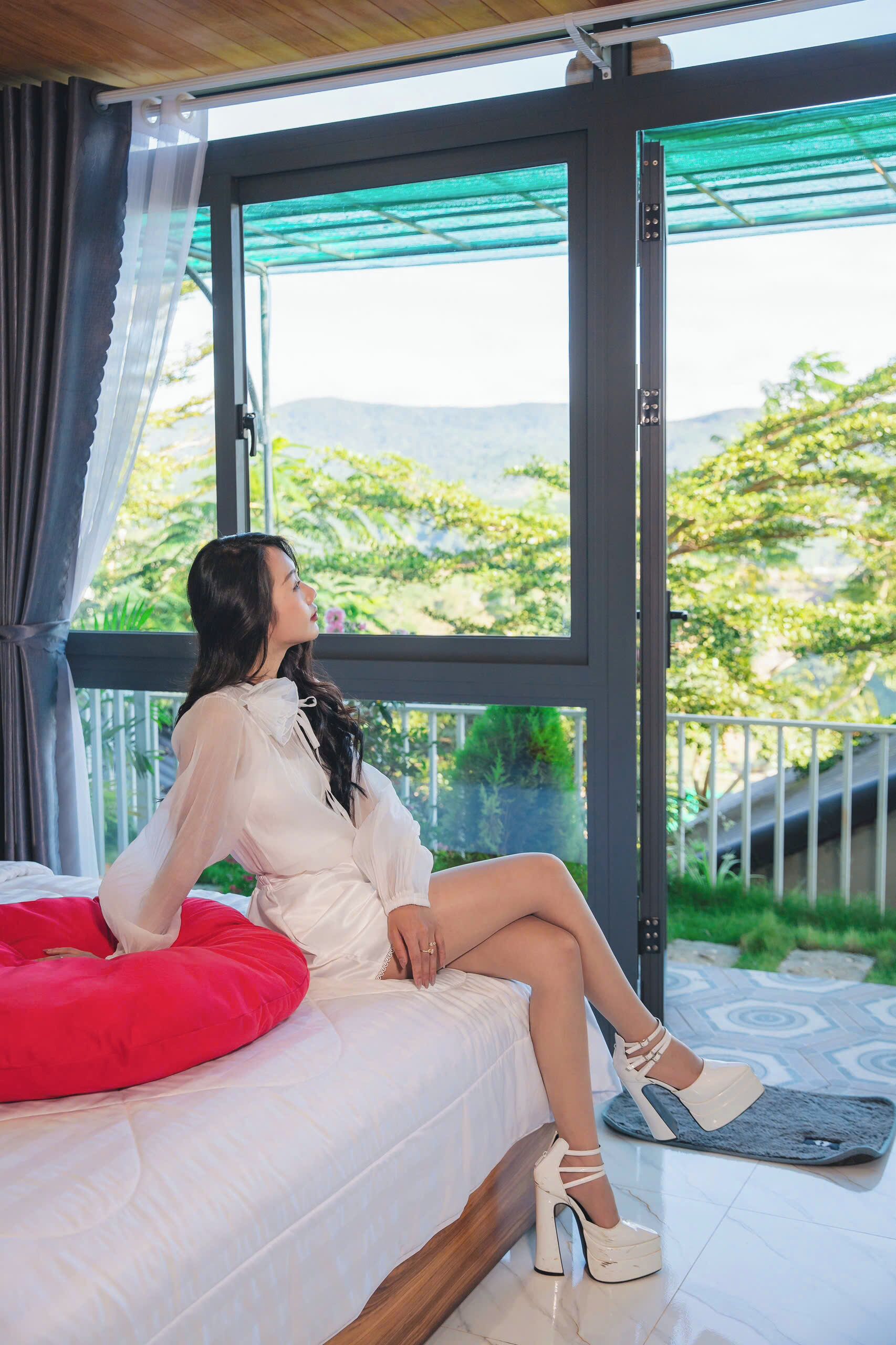 A woman sitting on a bed with a red pillow, wearing white high-heeled shoes, in a room with a large window overlooking greenery.