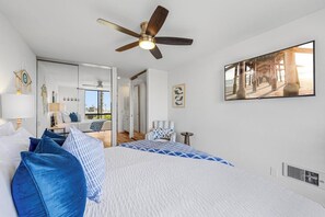 A bedroom with a bed, a ceiling fan, a TV, and a view of the beach through the window.