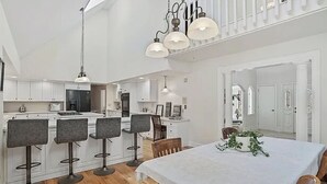 A modern kitchen with white cabinetry, a central island, and bar stools. There is a dining area with a table and chairs, and a staircase with a railing leading to an upper level. - Cheerful 6-Bedroom Villa with Pool and Tennis Courts near the beach! (Fairfield)