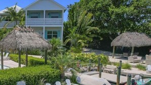A poolside area with lounge chairs, a thatched-roof structure, and a building in the background. - Isla Lychee: Boutique Resort, FREE Boat Dockage Sep-Oct Only! (Islamorada)