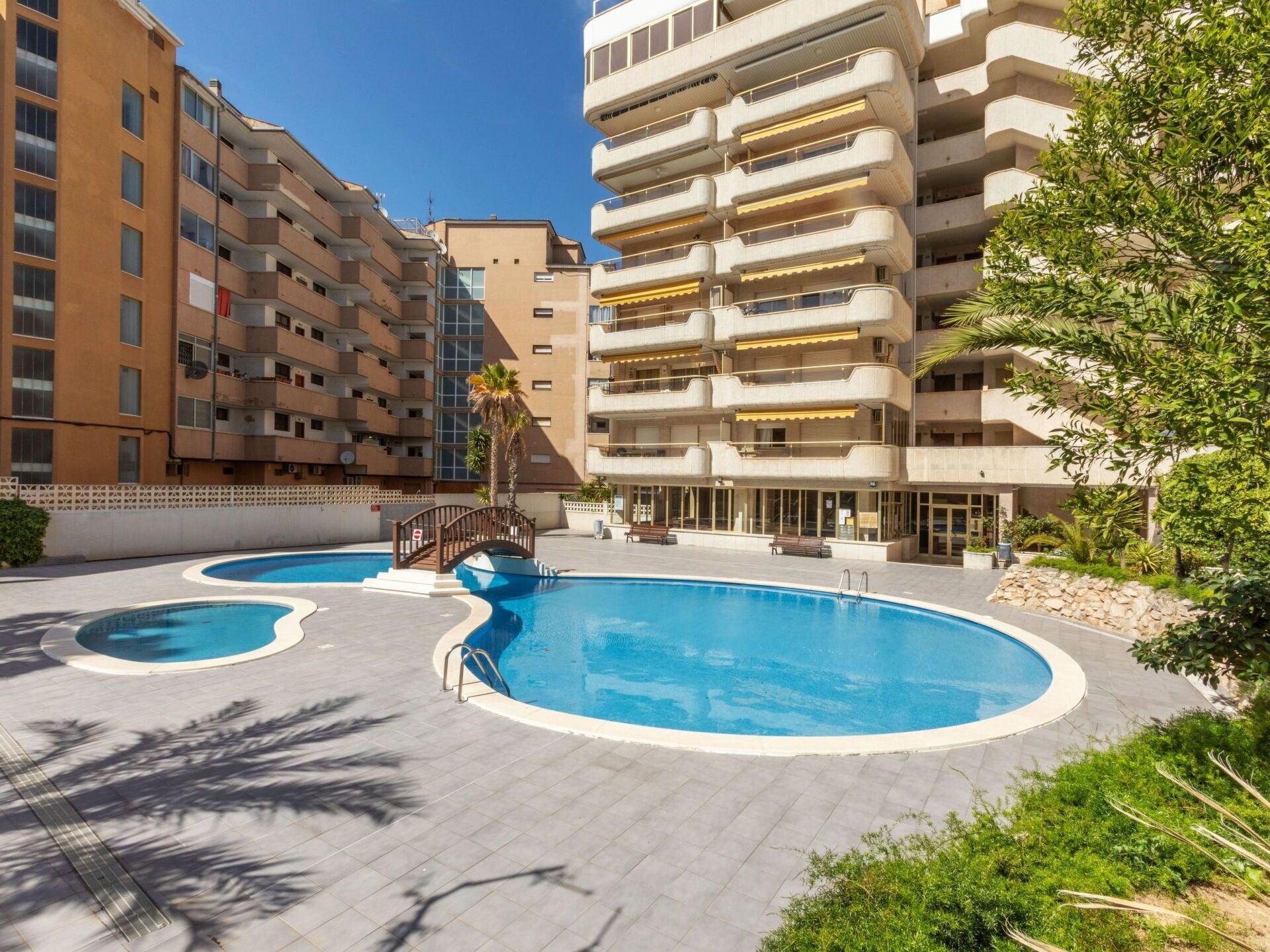 Zona de piscina con varias piscinas y un puente, rodeada de edificios de apartamentos.