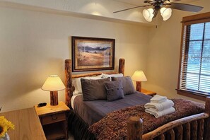 Un dormitorio con una cama, dos lámparas de noche, un ventilador de techo, un cuadro en la pared y una ventana con persianas.