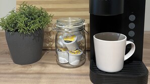 Eine Kaffeemaschine von Keurig, eine weiße Tasse, ein Glasbehälter mit Kaffeekapseln und ein Blumentopf auf einer Holzoberfläche.