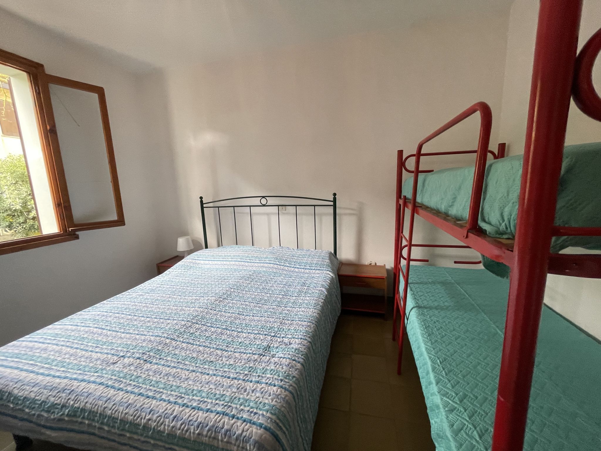 A single bed with a patterned bedspread, a window with a wooden frame, and a bunk bed with green bedding.