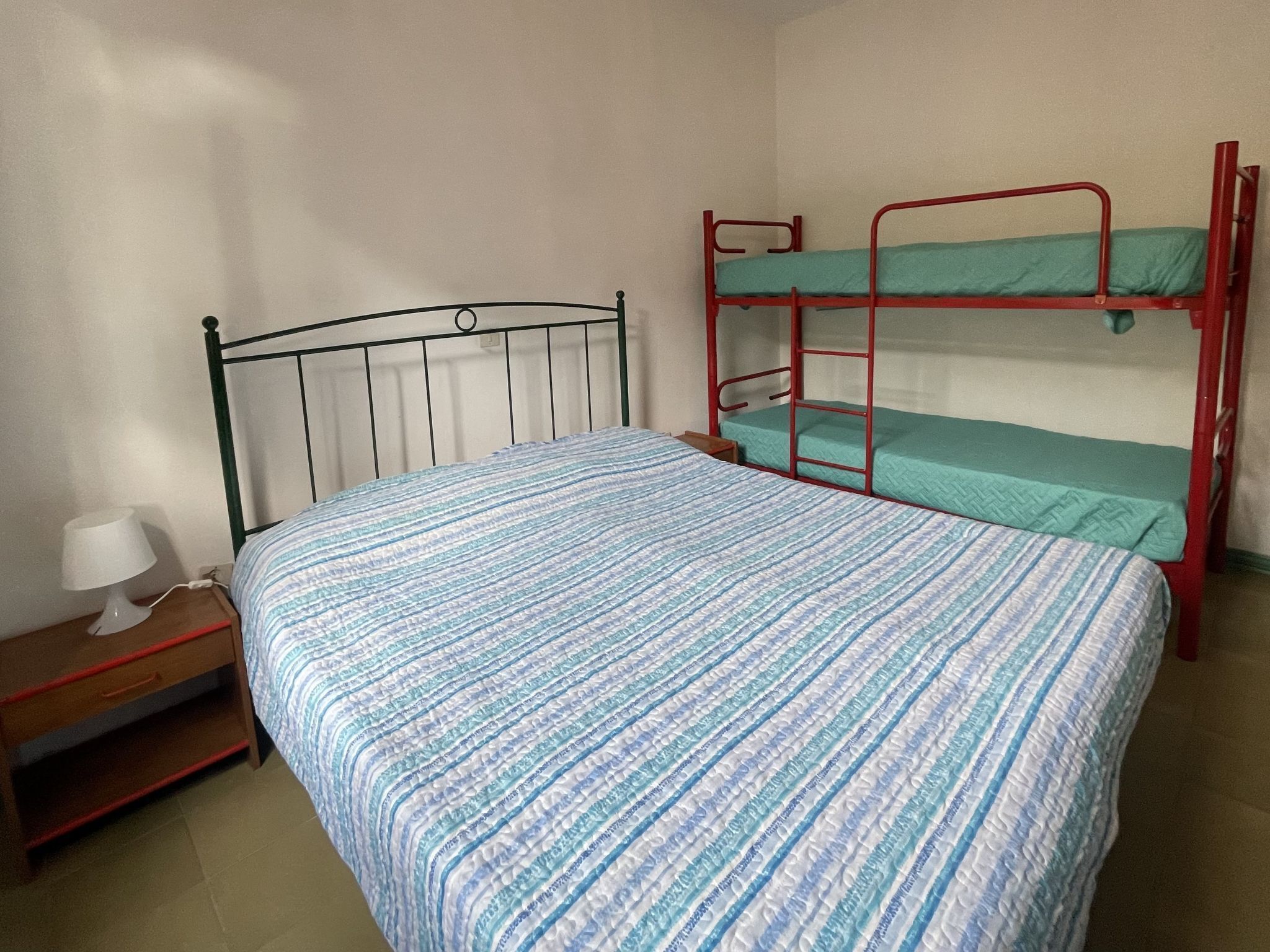 Ein Einzelbett mit blau-weiß gestreifter Bettdecke, ein Nachttisch mit Lampe und ein Stockbett mit grüner Bettwäsche.