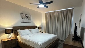 Une chambre à coucher avec un lit en bois, une table de chevet avec une lampe, un ventilateur de plafond et une fenêtre avec des rideaux.
