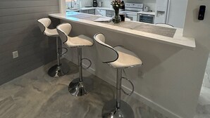A modern kitchen with grey cabinets, a stainless steel refrigerator, a microwave, and a built-in oven. There are three bar stools with chrome legs and white seats. - Beautiful Family-sized Home in Sunny Sarasota, Florida with large backyard! (Sarasota)