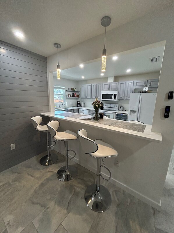 A modern kitchen with grey cabinets, a stainless steel refrigerator, a microwave, and a built-in oven. There are three bar stools with chrome legs and white seats. - Beautiful Family-sized Home in Sunny Sarasota, Florida with large backyard! (Sarasota)