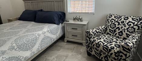 Une chambre à coucher comprenant un lit, une table de chevet, une chaise, une fenêtre avec des stores et deux œuvres d’art murales.