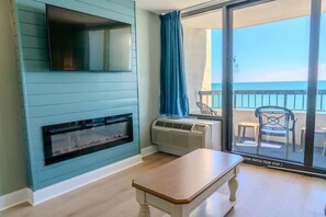 A room with a fireplace, a TV mounted on the wall, a table, and a view of the balcony with outdoor furniture.