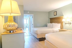 A hotel room with two beds, a lamp, a bedside table, and a view of the outside.