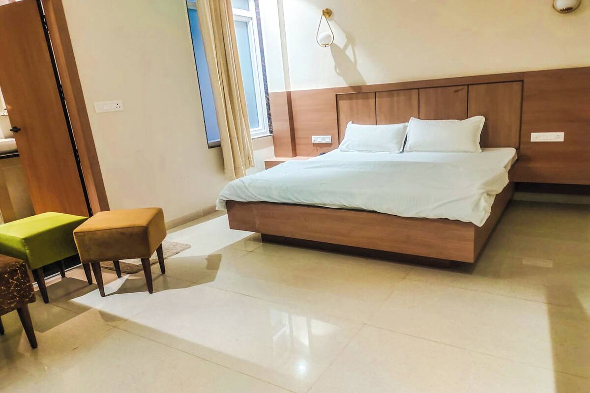 Een hotelkamer met een groot bed, twee kleine voetenbankjes, een houten hoofdbord en een raam met gordijnen.