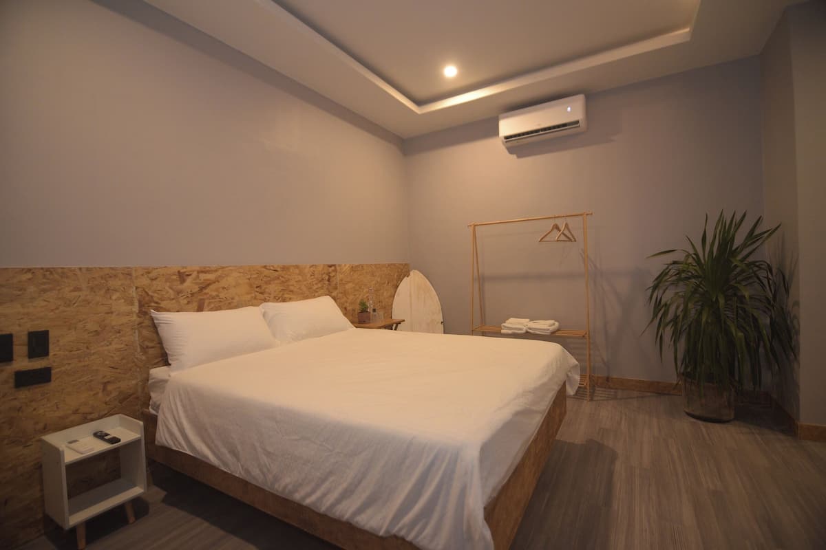 Une chambre à coucher avec une tête de lit en bois, un lit recouvert de linge de lit blanc, une table de chevet avec une lampe, un miroir et une plante en pot.
