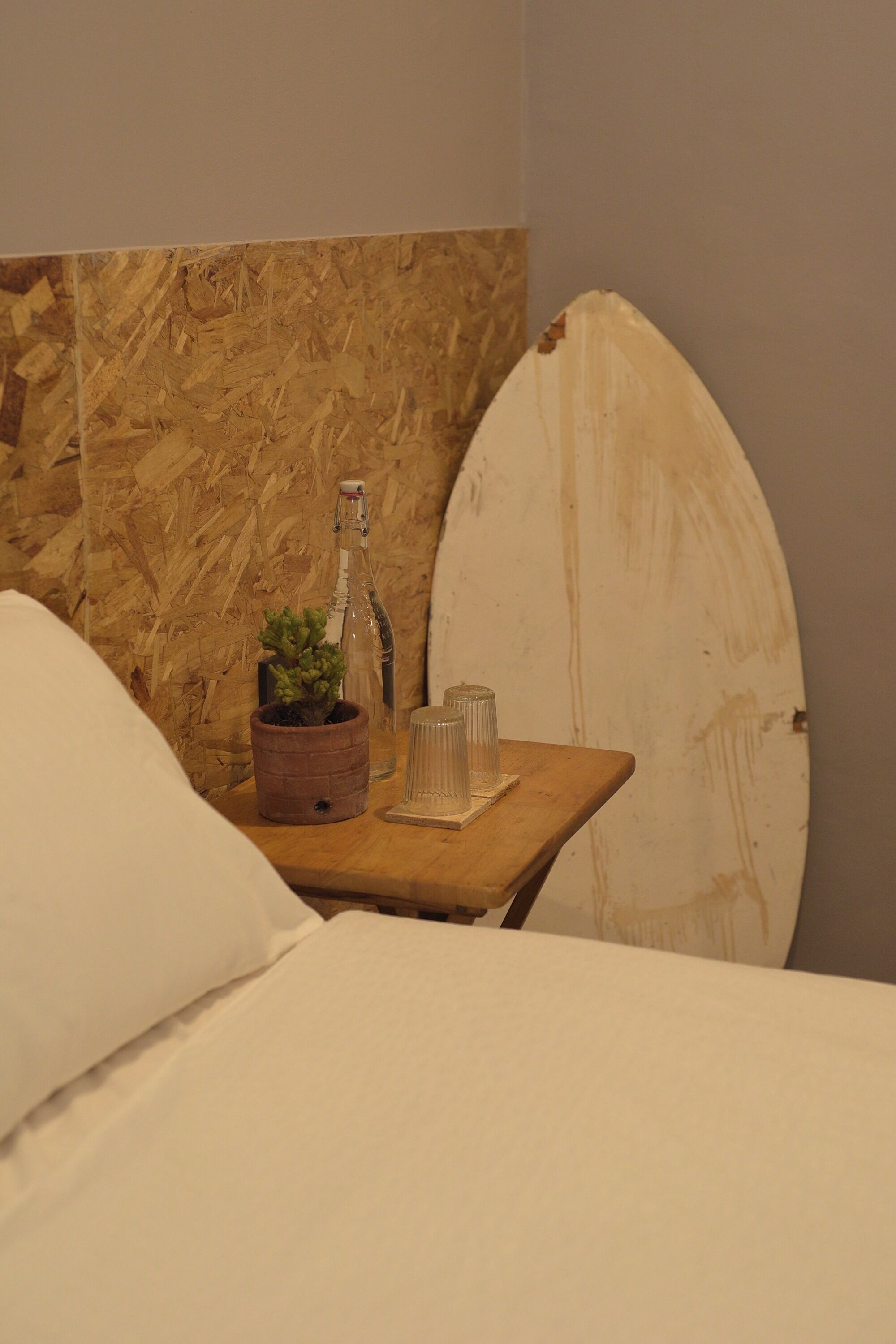A bed with white bedding, a small wooden table with a potted plant, a bottle, and a glass.