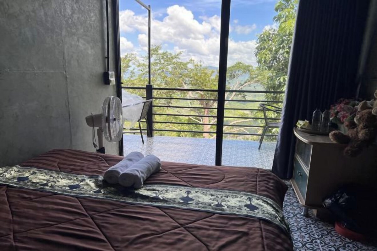 Une chambre avec un lit, un ventilateur, une fenêtre et une vue sur la verdure.