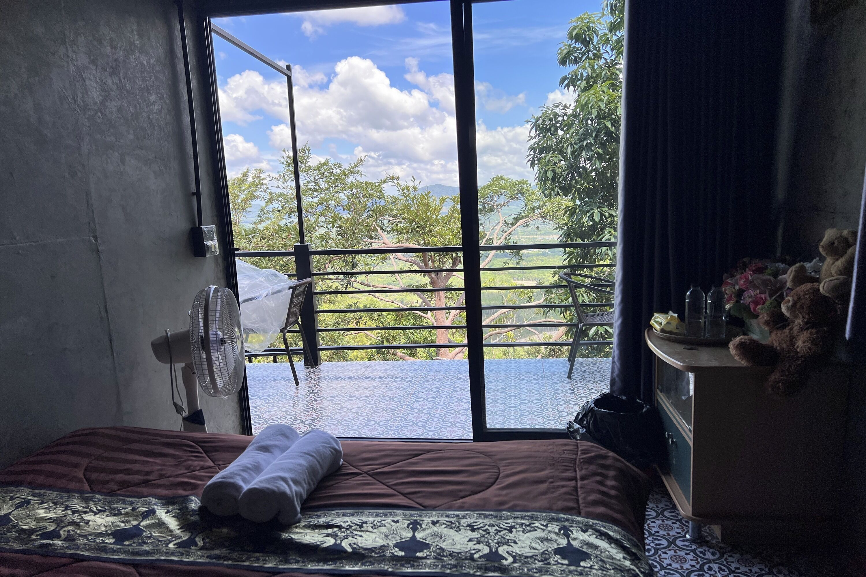 Une chambre avec un lit, un ventilateur, une commode et une vue sur la verdure.