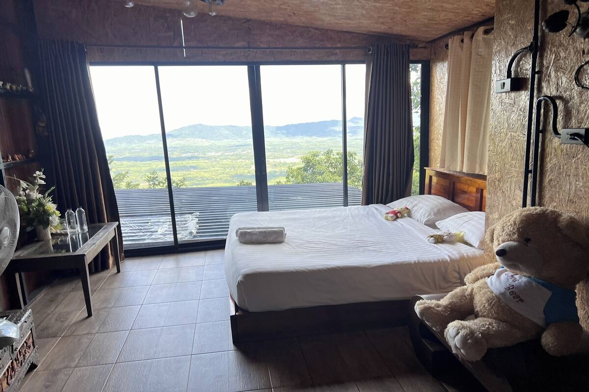 Een slaapkamer met een groot bed, een teddybeer, een ventilator en uitzicht op de omgeving.