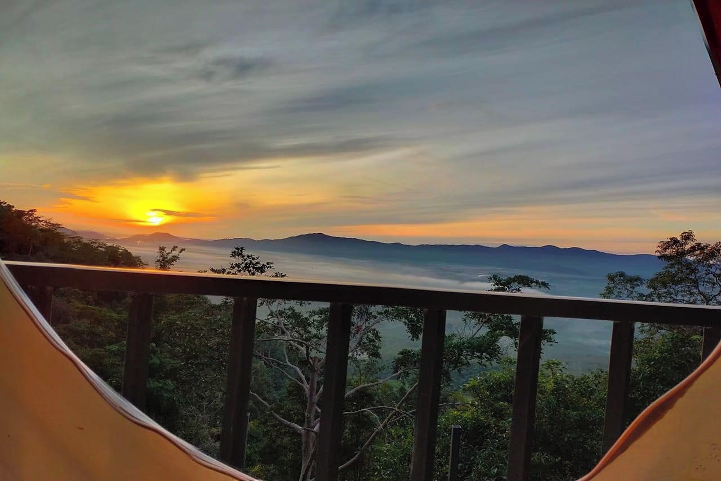 A sunset view from a balcony with a railing, overlooking a mountainous landscape and dense forest.