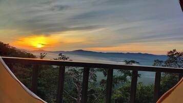 A sunset view from a balcony with a railing, overlooking a mountainous landscape and dense forest.