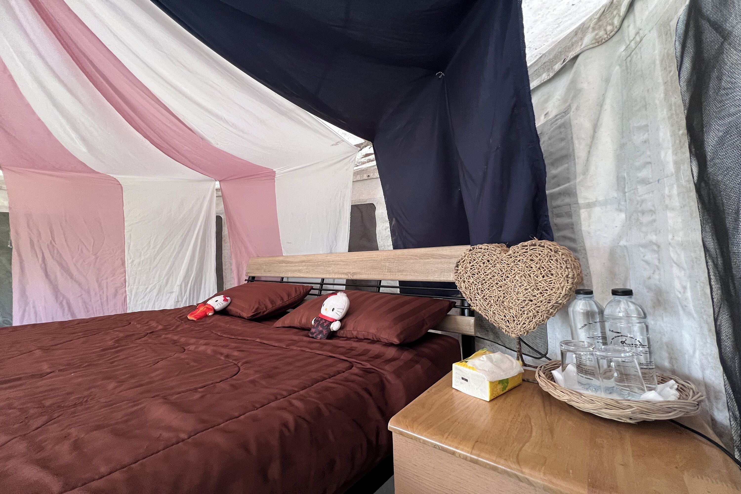 Un lit recouvert d’une couverture marron, un doudou à l’effigie de Hello Kitty et un oreiller en forme de cœur. Sur la table de chevet, on trouve une boîte de mouchoirs et des lunettes.
