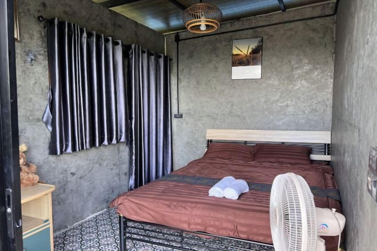 Une chambre à coucher de taille réduite, avec un lit simple, un ventilateur de chevet et une petite table avec une lampe.