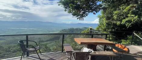 A wooden deck with a table and chairs, overlooking a scenic landscape with mountains and greenery.