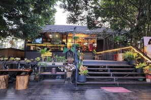 An outdoor seating area with wooden tables and chairs, surrounded by potted plants and a staircase leading to an upper level. - Kuan Khan Lao The Nine Bistro (Na Mom)