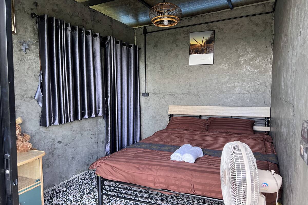 Een slaapkamer met een bed, een ventilator, een teddybeer en een foto aan de muur.