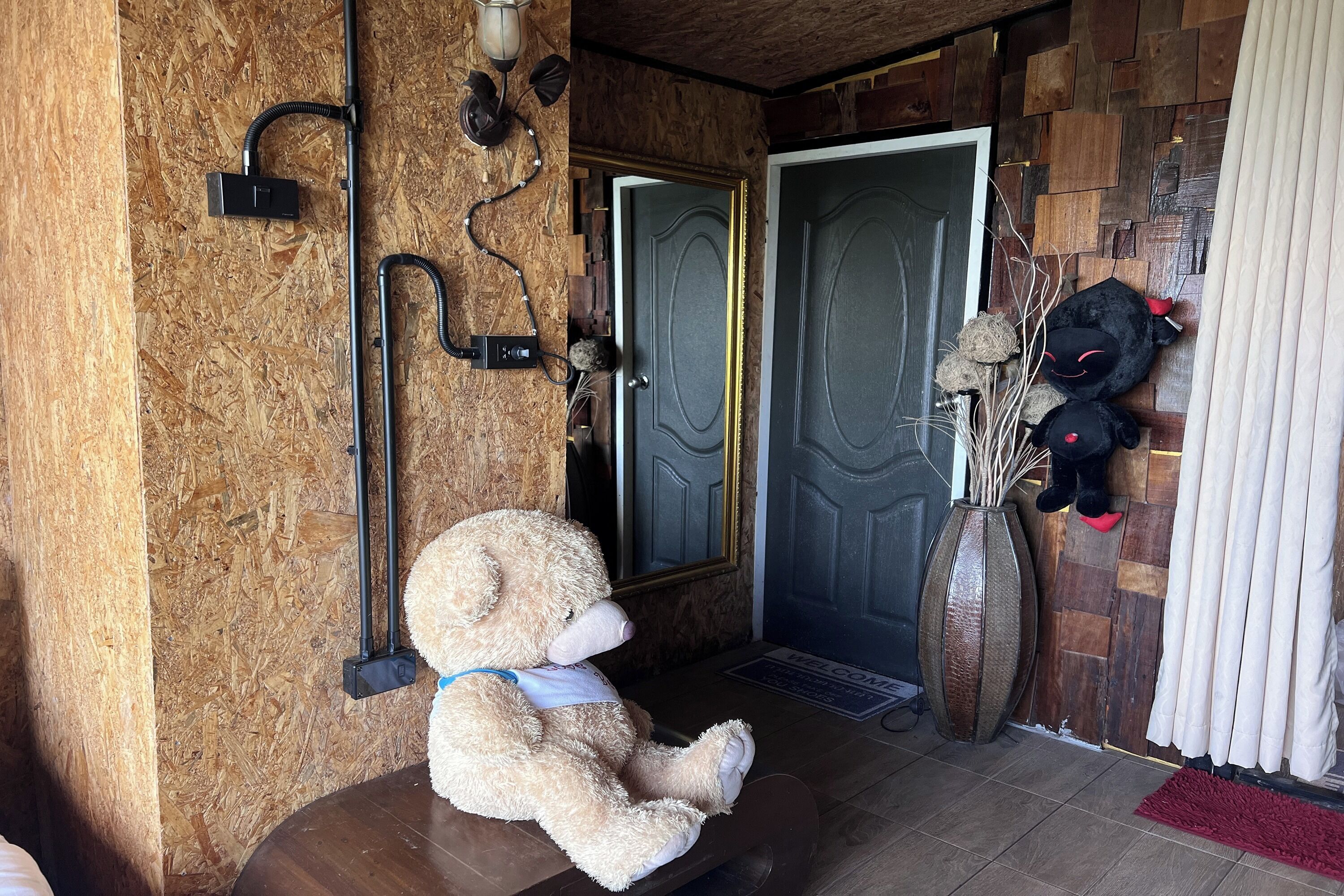 Une pièce avec un mur en bois, une porte, un grand vase rempli de branches et un ours en peluche posé sur un banc.