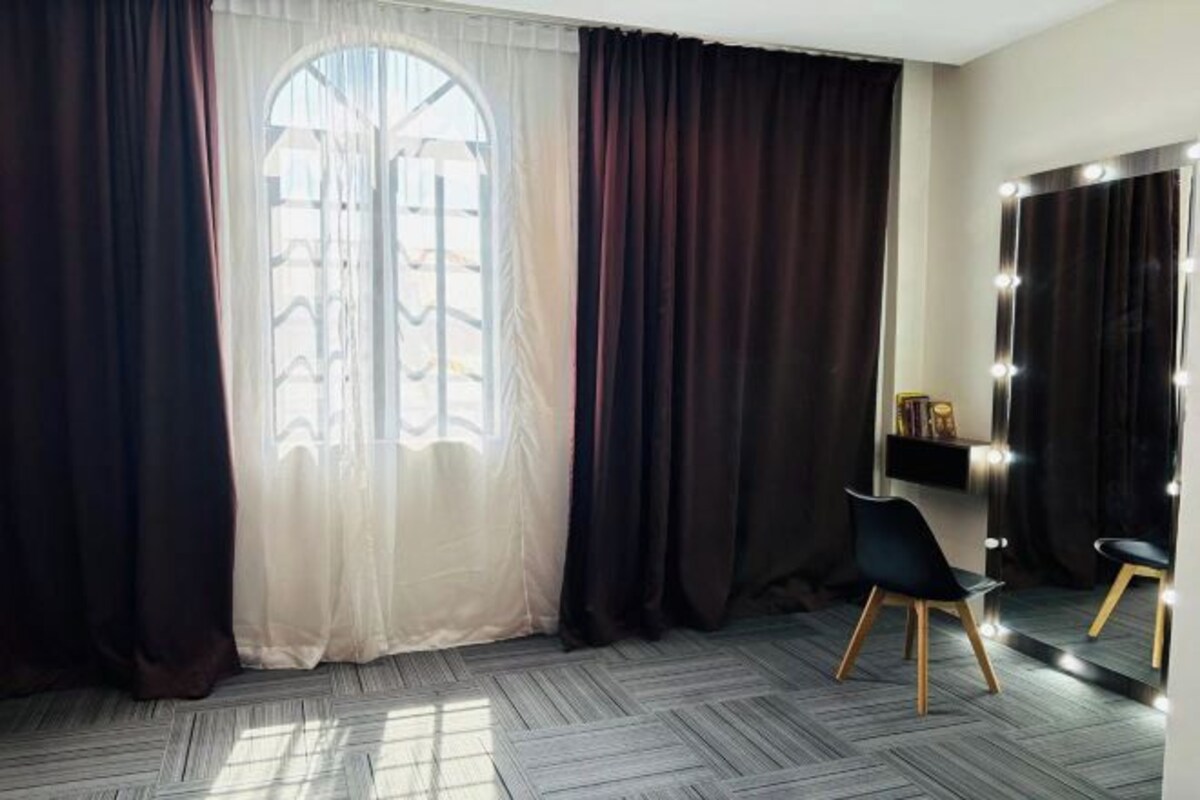 A room with a large window, dark curtains, a mirror with lights, a chair, and a shelf with books.