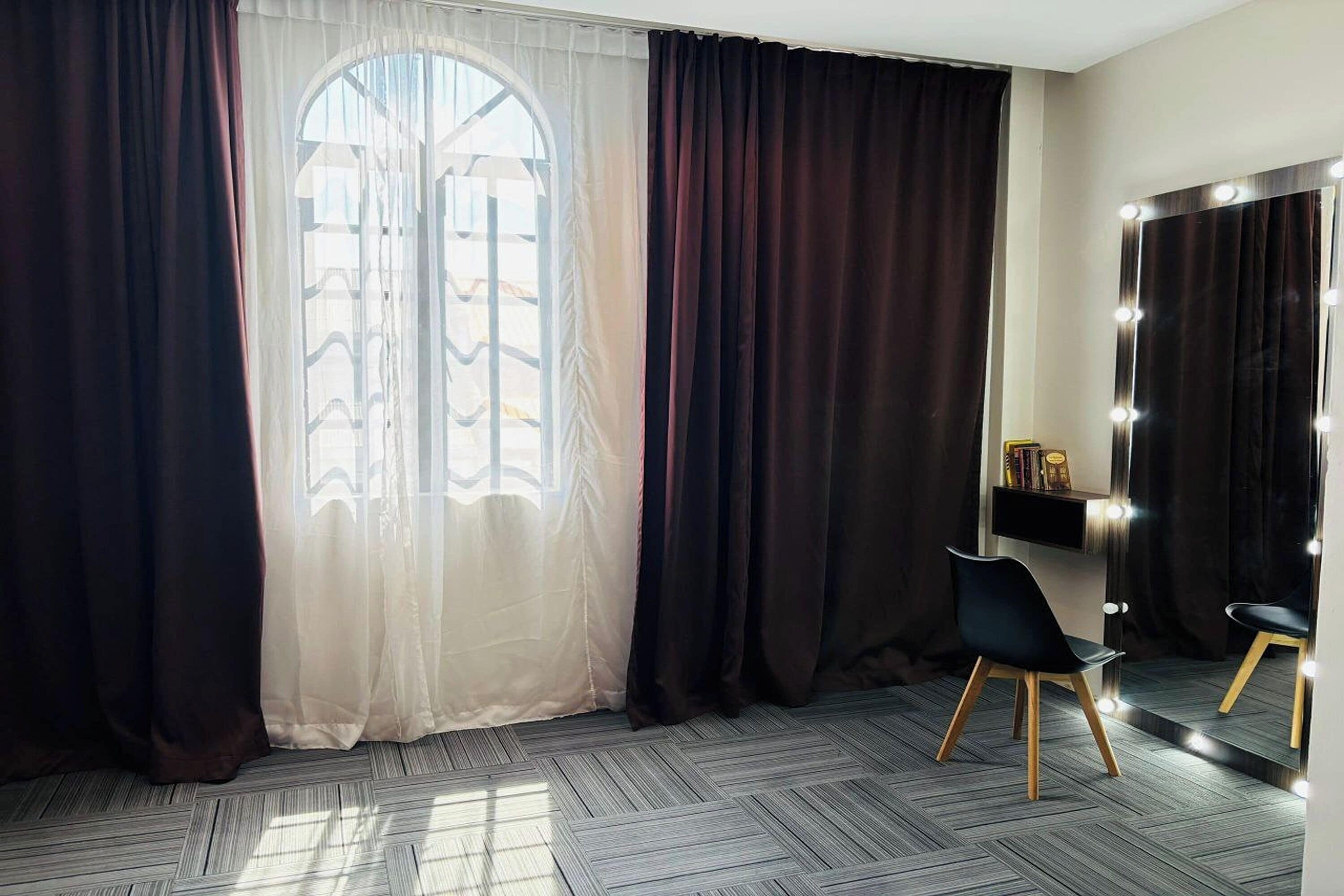 A room with a large window, dark curtains, a vanity with lights, a chair, and a herringbone-patterned floor.