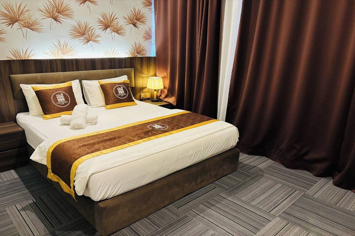 Una habitación de hotel con una cama grande, ropa de cama marrón y blanca, un cabecero, una mesita de noche con una lámpara y una ventana con cortinas.