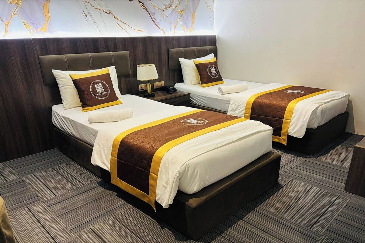 Habitación de hotel con dos camas, cada una con cabecero marrón, ropa de cama blanca y un corredor de cama marrón y amarillo. Hay una mesita de noche con una lámpara y un teléfono entre las camas.