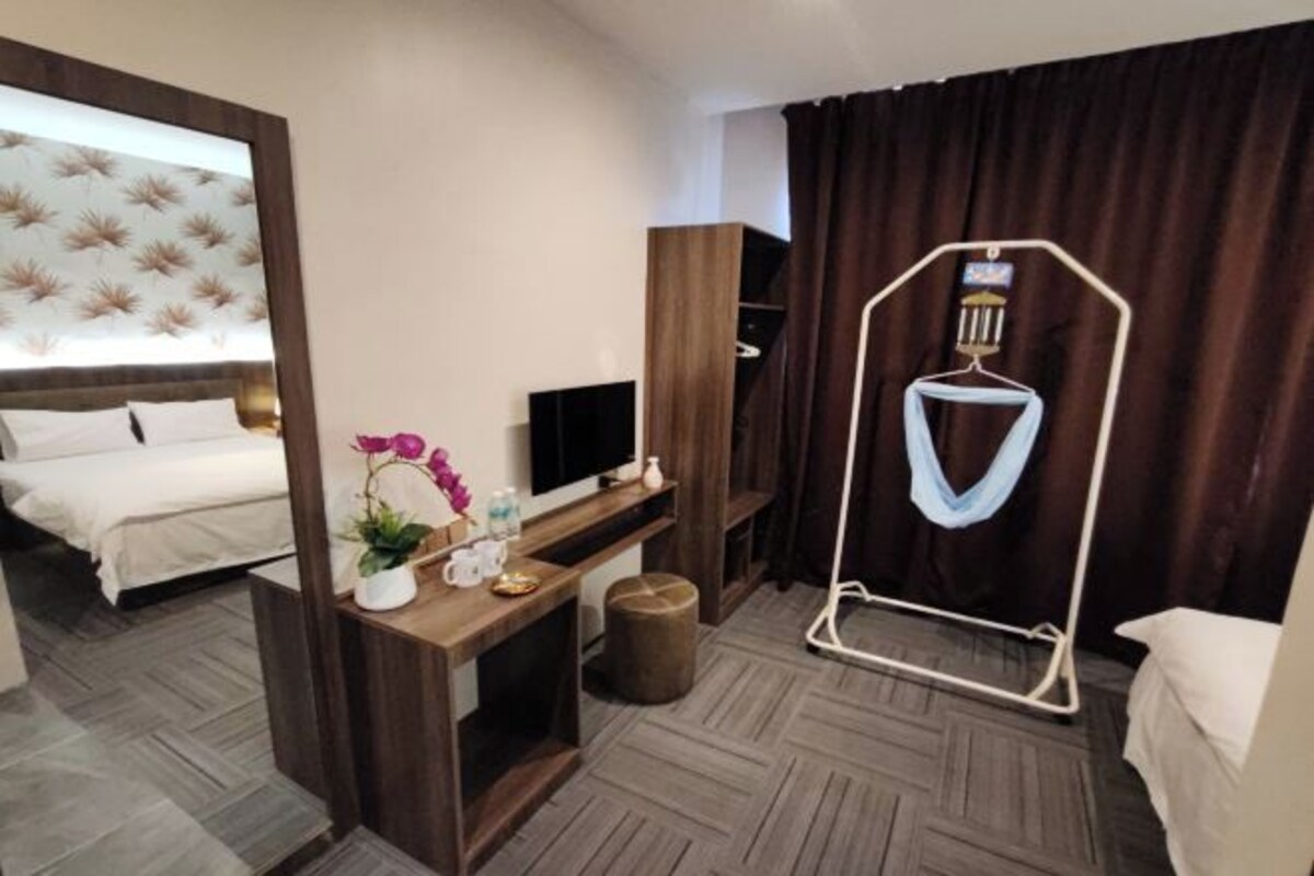 A hotel room with a bed, a desk with a TV, a small plant, and a hanging light fixture.