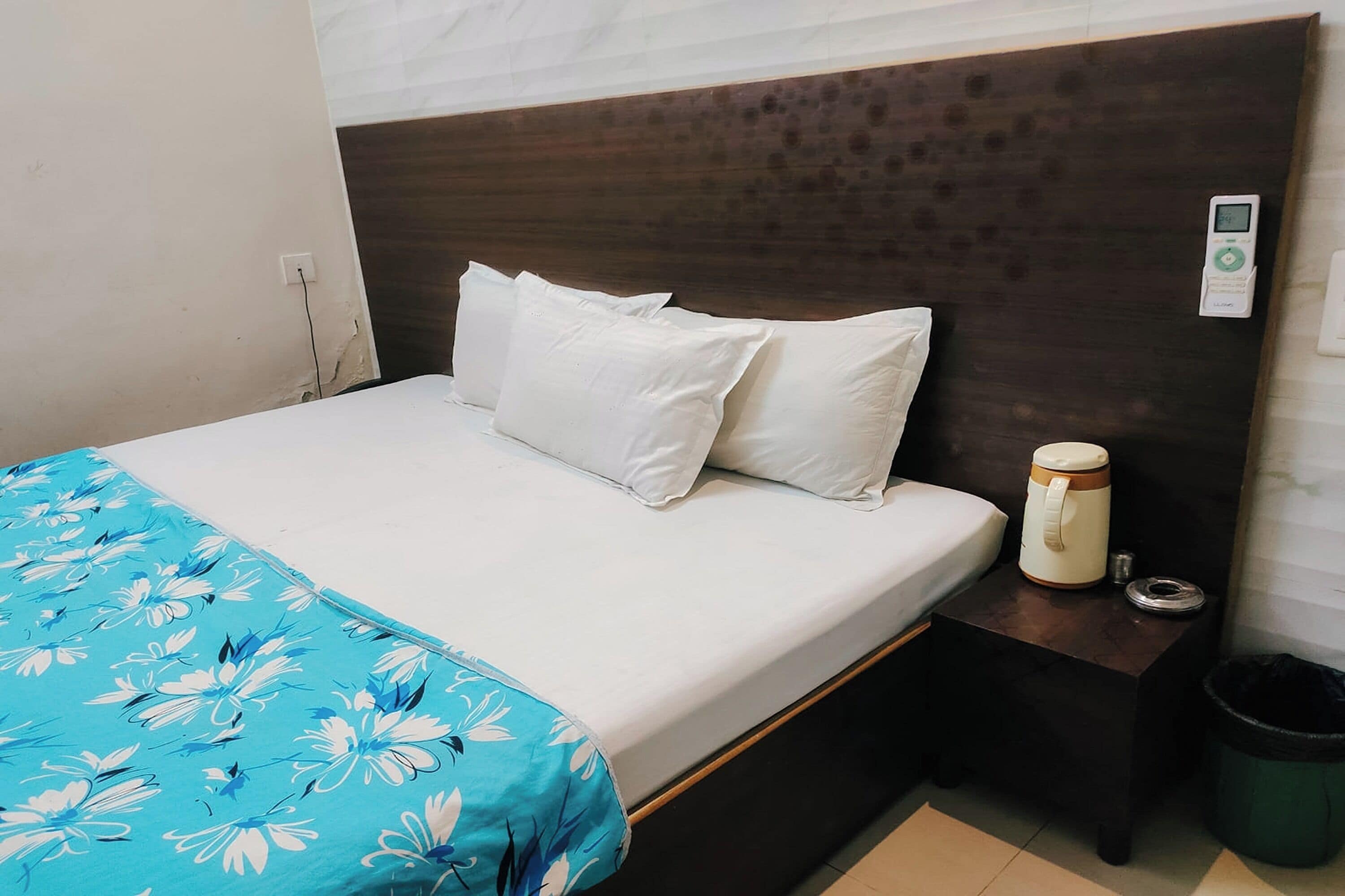 Una cama con ropa de cama blanca y un plaid floral azul, una mesita de noche con cafetera y un cubo de basura.