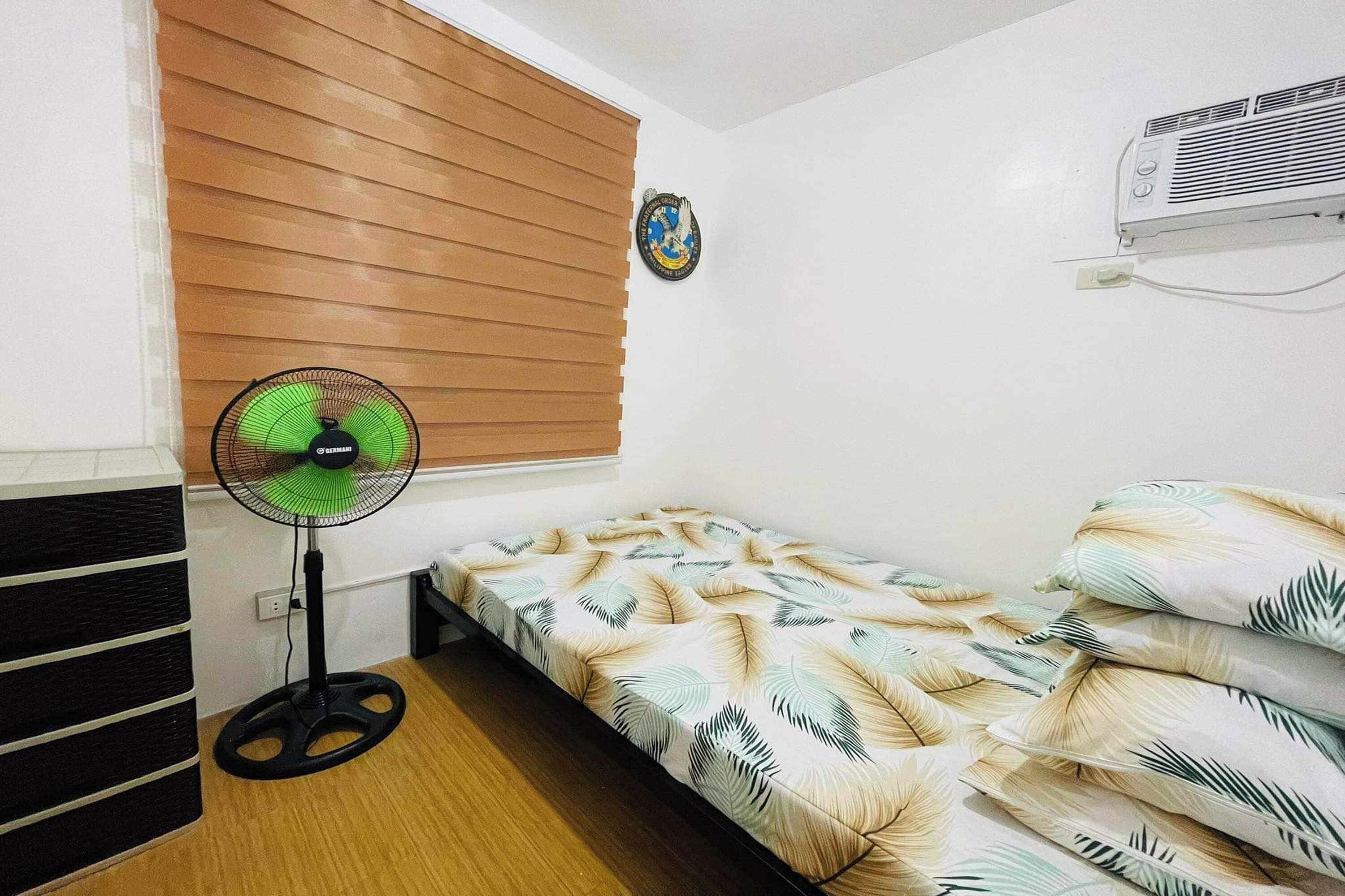 Una habitación pequeña con cama, ventilador de pie, aire acondicionado instalado en la pared y una ventana con persianas.