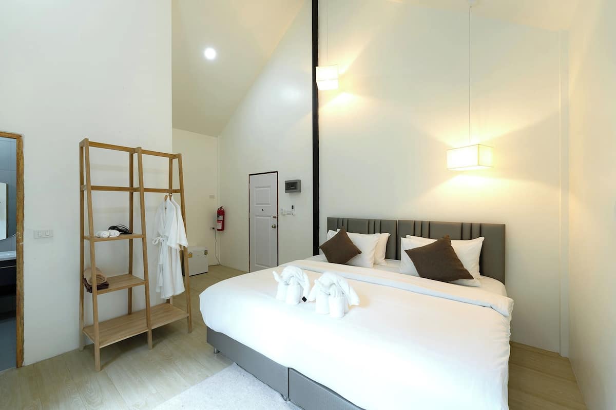 A modern hotel room with a large bed, a wooden shelf with towels, a door, and a wall-mounted light fixture.