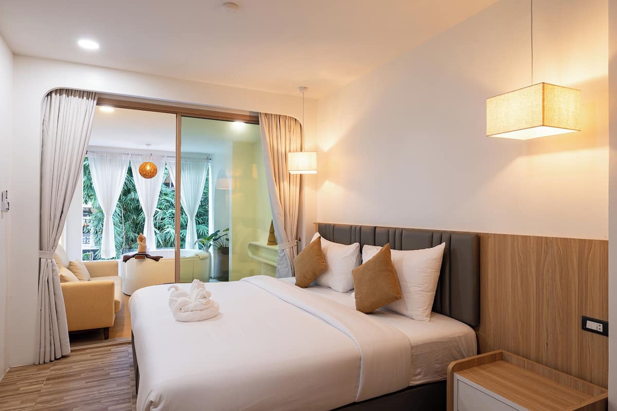 A modern hotel room with a large bed, a bedside table, a chair, and a view of greenery through the window.