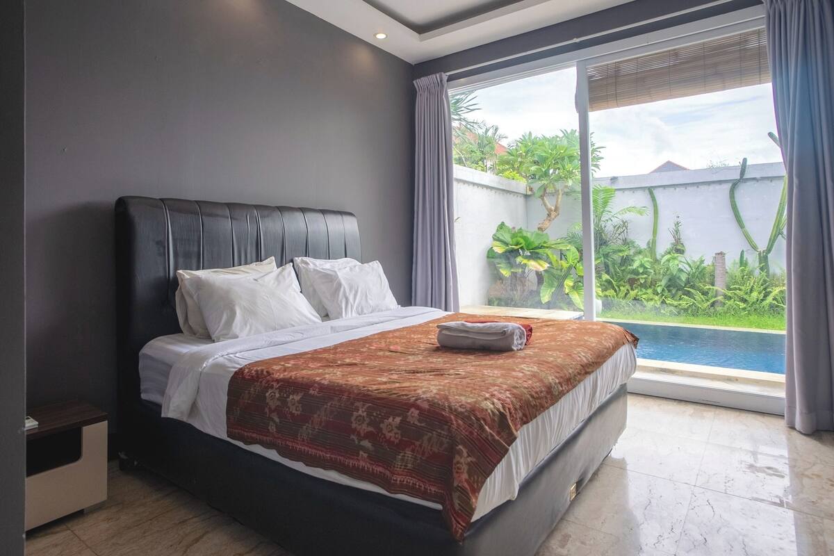 Une chambre avec un grand lit, une fenêtre donnant sur une piscine et un espace verdoyant, ainsi qu’une table de chevet.