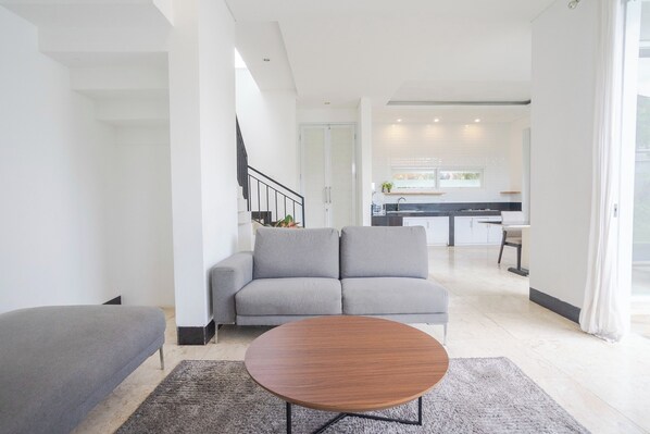 A modern living room with a grey sofa, a round wooden coffee table, and a staircase leading to an upper floor. - Villa And Surf Hostel Blue Margarita (Canggu)