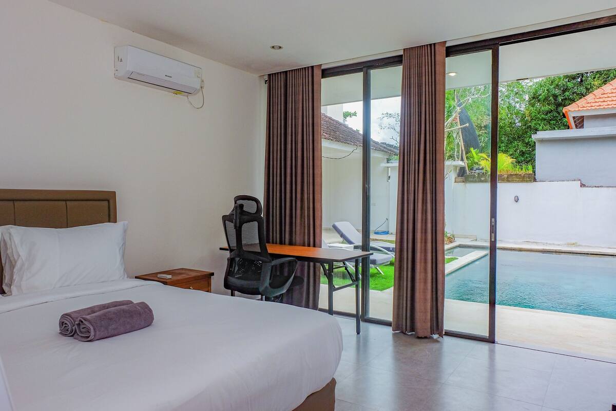 Une chambre d’hôtel comprenant un lit, un bureau, une chaise, une fenêtre avec des rideaux et donnant sur une piscine.