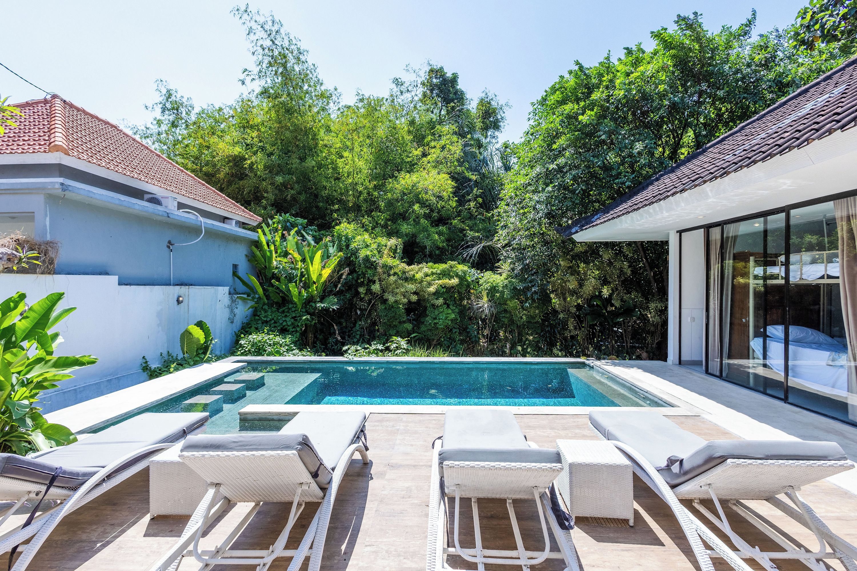 A swimming pool with lounge chairs, surrounded by lush greenery and a modern house with a tiled roof.