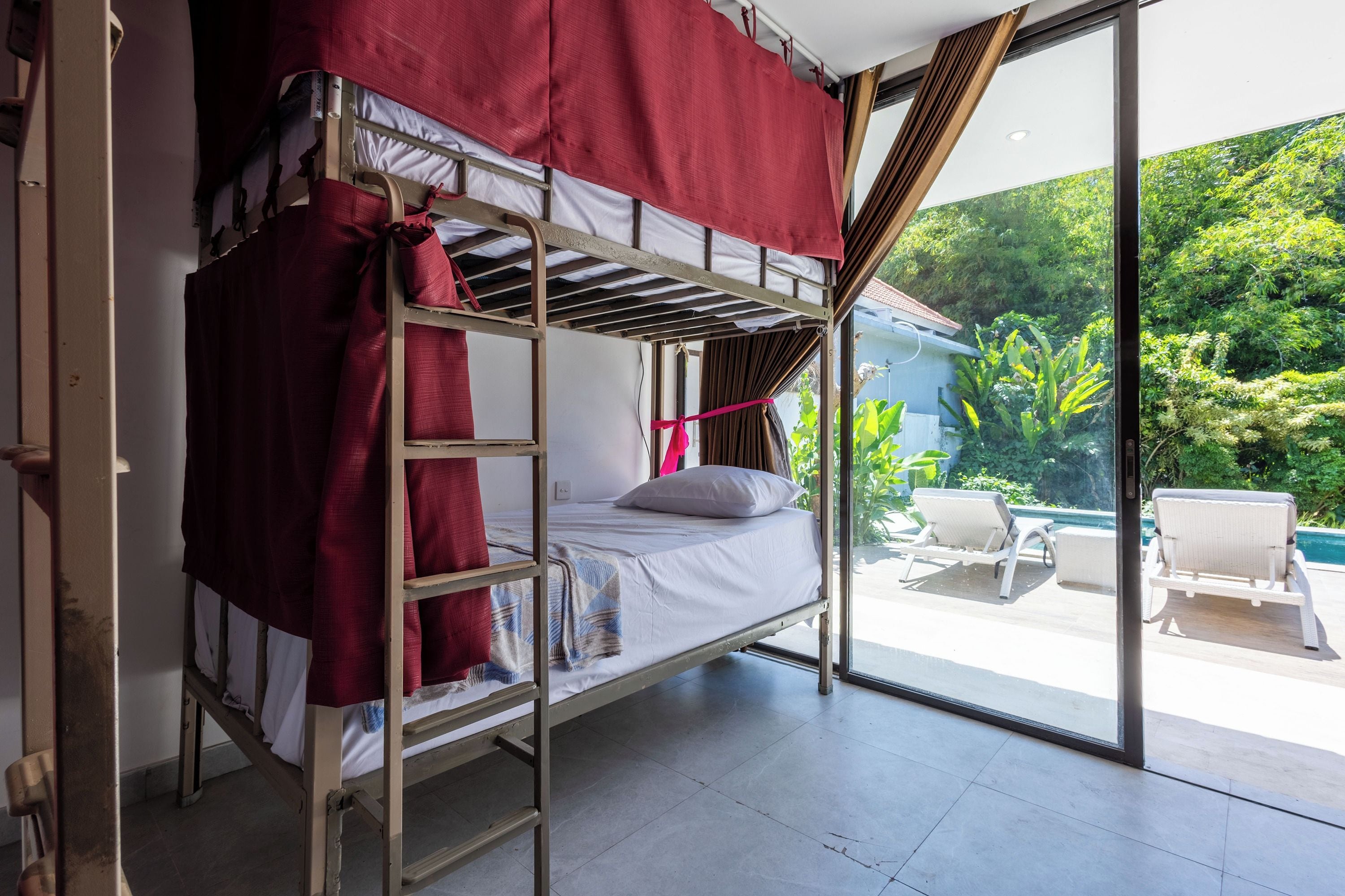 Un lit superposé avec une couverture rouge et un lit simple avec un oreiller, une porte vitrée donnant sur un espace extérieur avec une piscine et des chaises longues.
