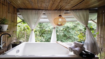 A bathroom with a large sink, white curtains, a hanging light fixture, and a view of greenery outside.