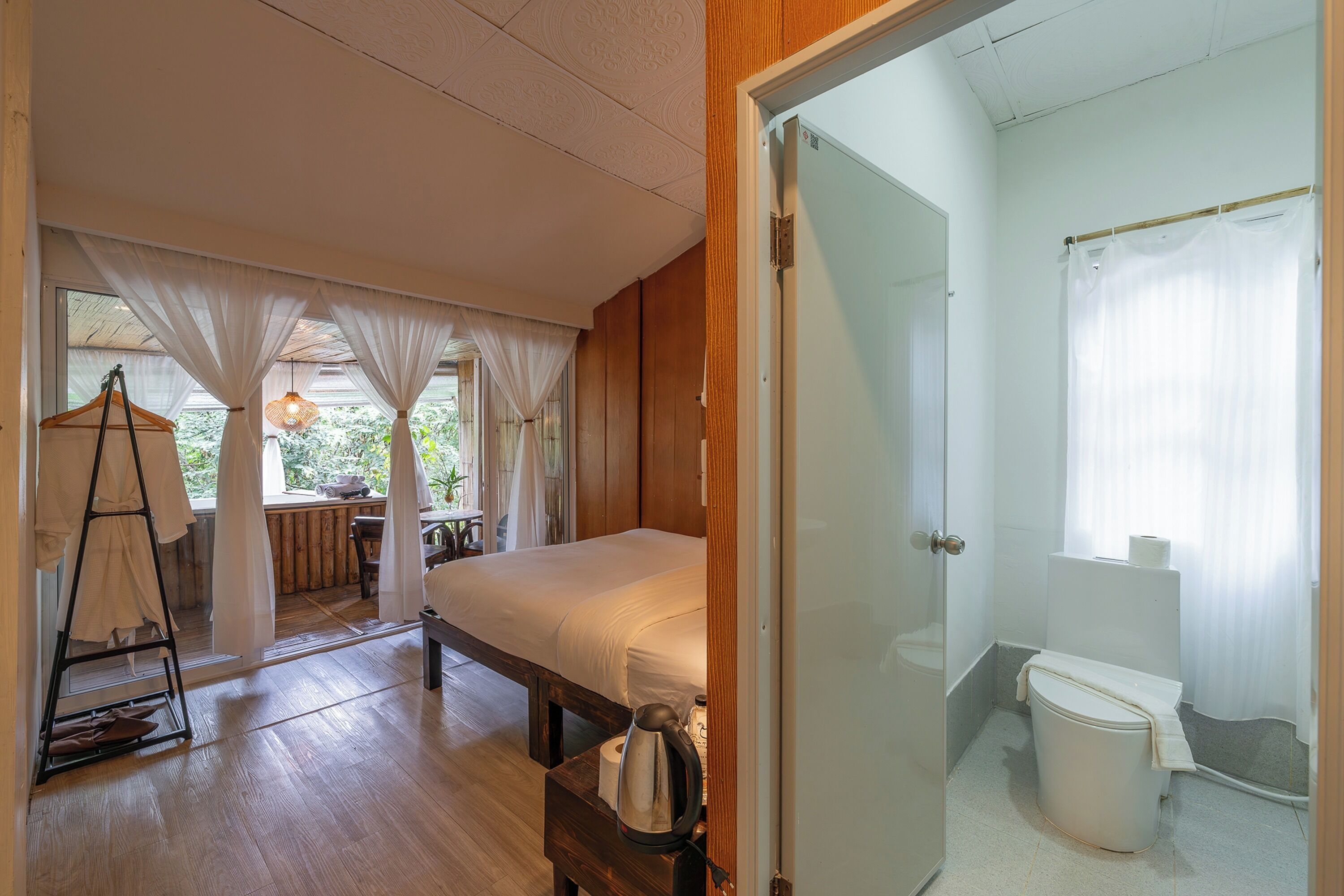 Camera da letto con pavimento in legno, un letto, un comodino con bollitore e vista su un balcone con tende.