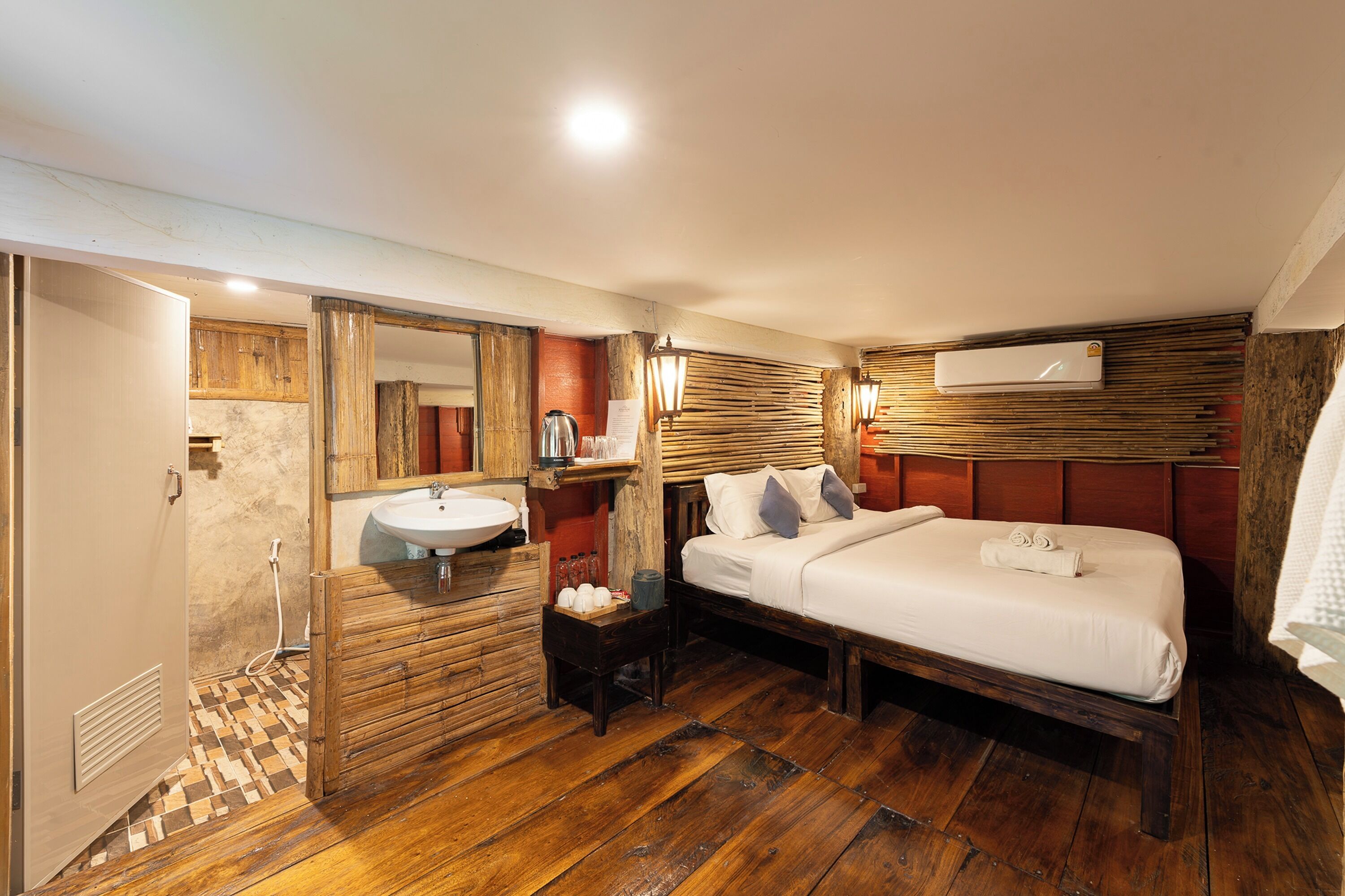 A hotel room with a wooden floor, a bed with white linens, a wooden vanity with a sink, a mirror, and a small table with a kettle and cups.