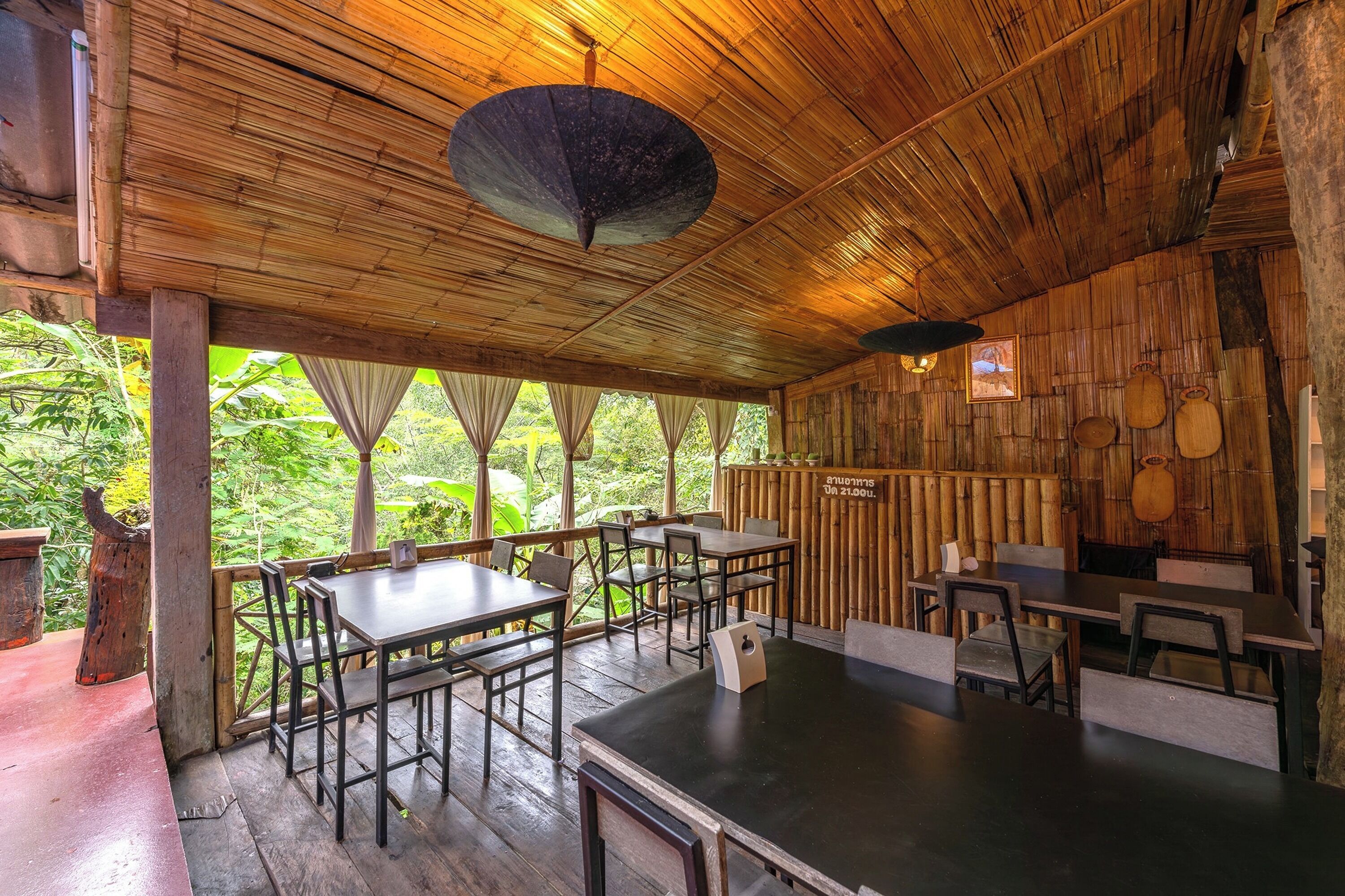 An indoor dining area with wooden tables and chairs, a bamboo ceiling, and a view of greenery outside.