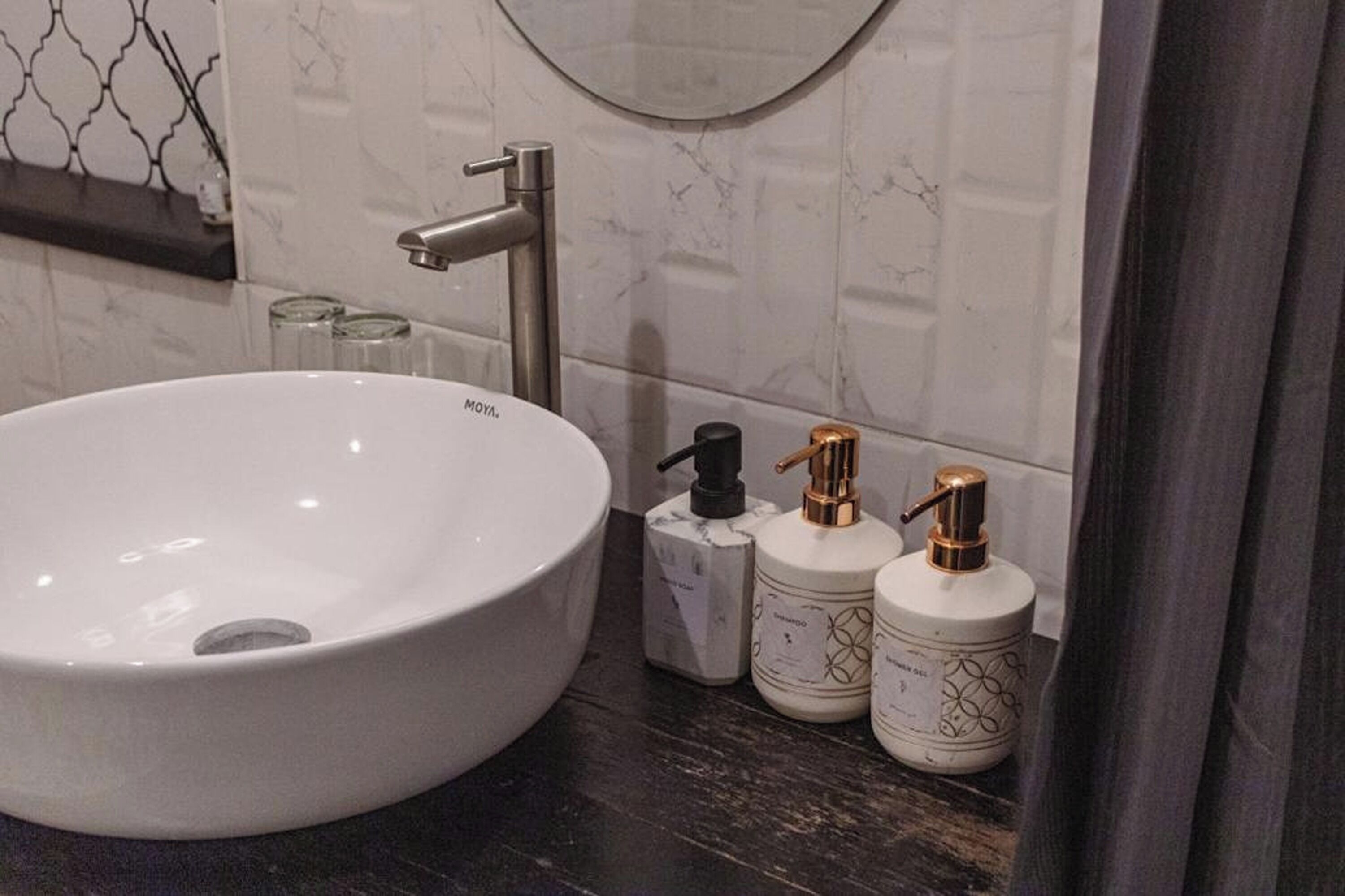 Une salle de bain avec un lavabo sur colonne blanc, un robinet moderne et deux flacons de produits de toilette ornés de détails dorés, le tout posé sur un plan de toilette foncé.